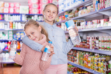 Portrait of cheerful little girls standing among shelves in the supermarket, holding a bottled water