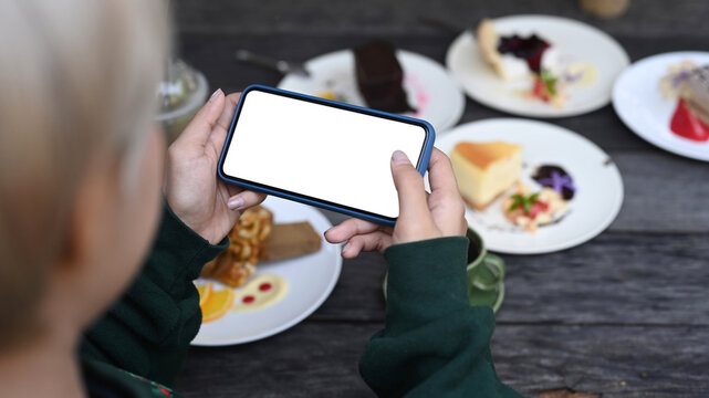 Young Asian girl holding horizontal smart phone taking photo of sweet desserts at cafe.