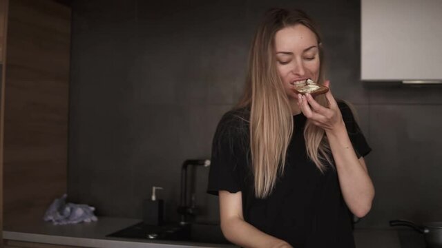 Blonde woman eating bread with butter for breakfast, checking her smartphone in kitchen