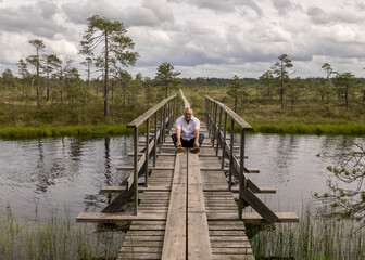 the summer swamp. a man in a white shirt sits on a wooden bridge. bog pond. bog background and vegetation