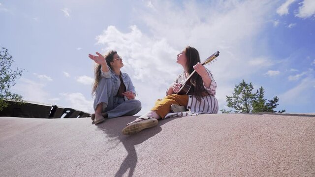 Two young woman sitting on concrete playground under high cloudy sky singing and playing ukulele guitar while relax in modern city park enjoying sunny summer day. low angle shot 