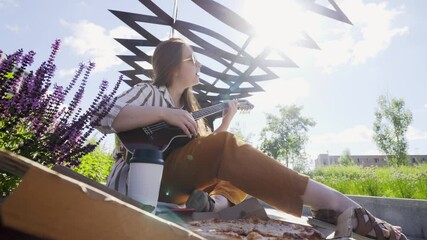 happy young friends picnic on bench in contemporary city park on sunny summer day. Lady playing on ukulele and eating slices of delicious cheese pizza from box in bright sunny backlit - Powered by Adobe