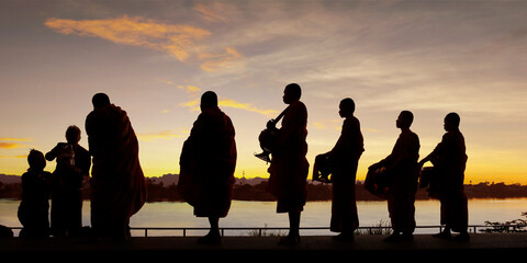 silhouette of monks dressing orange robe during reception of alms, on the shore of the mekong river
