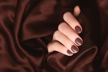 Female hand with brown nail design. Brown nail polish manicure. Woman hand on dark brown fabric background.