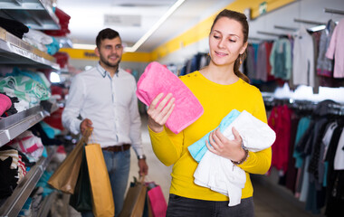 Naklejka na ściany i meble Glad girl buying towels in shop while her boyfriend standing behind with purchases