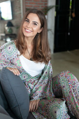 attractive brunette smiling girl with long hair in white top and silk trouser suit with indian pastel multicolor paisley pattern sits on gray sofa in interior