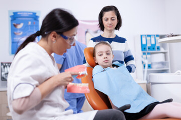 Ortodontist brushing artificial jaw educating child sitting on dental chair. Little girl and mother listening stomatolog talking about tooth hygine in dentistiry clinic holding jaw model.