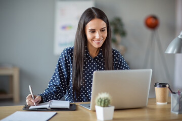 Photo of attractive cheerful young business woman write notebook look computer sit desk chief leader indoors in office