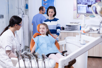 Dentist in dental office telling little girl the treatment will not be painfull. Child with her mother during teeth check up with stomatolog sitting on chair.