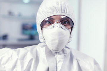 Exhausted microbiologist in coverall sitting in laboratory looking at camera. Tired woman scientist in biotechnology laboratory wearing protective suit during global epidemic.