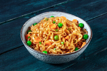 Instant noodles with vegetables in a bowl, with green peas and carrots on a dark blue wooden background