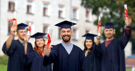 education, graduation and school concept - happy smiling male graduate student in mortar board and bachelor gown with diploma over group of people on campus background