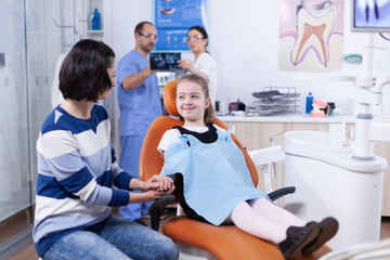 Happy little girl wearing dental bib in dentist office holding parent hand waiting tooth examination. Child with her mother during teeth check up with stomatolog sitting on chair.