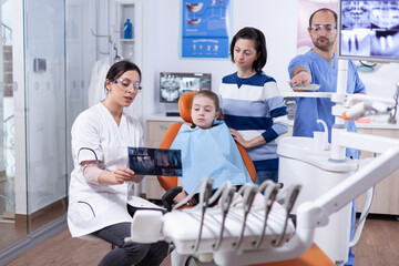 Dentist talking about tooth procedure holds radiography talking with little kid. Stomatologist explaining teeth diagnosis to mother of child in health clinic holding x-ray.