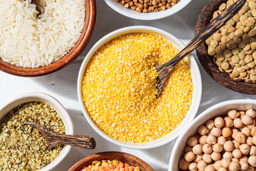 Raw corn grits in white bowl. Various dry cereals and legumes in white and wooden bowls, white wooden background. Vegan protein concept.