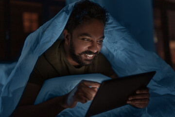 technology, internet, communication and people concept - young indian man with tablet pc computer lying in bed under blanket at home at night