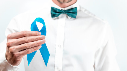Blue ribbon cancer. Awareness prostate of health in November. Hipster men in bright shirt, cyan bowtie with blue ribbon in hands on white background. Symbol of oncology affected man. Copy space.