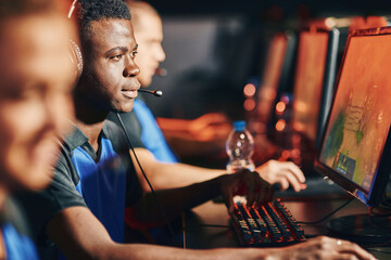 Young african guy, professional cybersport gamer wearing headphones playing online video game while sitting in a row with other team members in front of PC monitors © Friends Stock