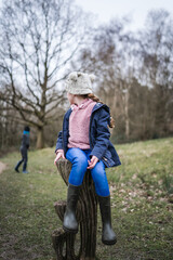 Fototapeta premium Excited happy young girl in wellinton boots sat in autumn countryside looking back away from camera at boy exploring the great outdoors nature park with bobble hat pink jumper and blue coat in winter