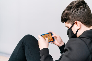Selective focus. A young teenager wearing a covid-19 mask plays with a smartphone online sitting on the street.