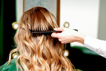Fototapeta premium Back view of hairdresser combing wavy hair of a young blonde woman in a beauty salon
