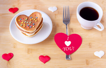 Homemade pancakes in the shape of a heart on a white plate, a white mug with coffee, a fork and a red paper heart with the inscription I LOVE YOU on a wooden background with hearts