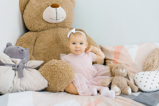 A cute one year old baby sits surrounded by soft animals toys and pillows