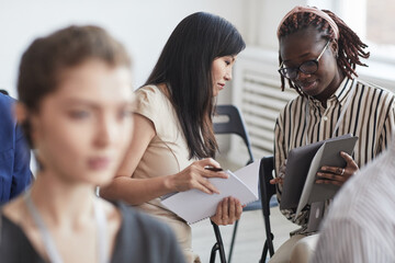 Portrait of two young women talking while sitting in audience at business conference or seminar,...