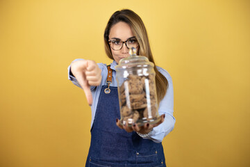 Young hispanic woman wearing baker uniform holding a cookies jar with angry face, negative sign showing dislike with thumbs down