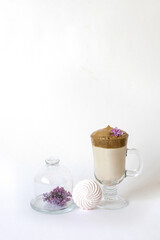 Dalgona coffee in a transparent mug with a lilac flower on a white background and a sprig of lilac lying on a serving plate and marshmallows. Spring or summer drinks menu