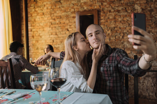 Man and woman in love take pictures together in a restaurant - An attractive Caucasian kisses her boyfriend's cheek while holding a smartphone for a selfie.