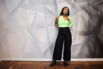 Fashionable african american woman in a light green top and black pants pose against grey modern wall.