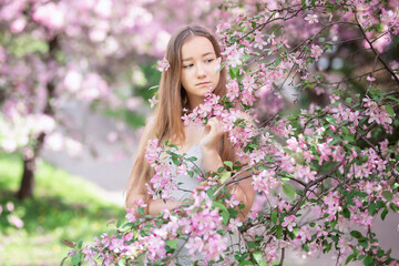 Fototapeta premium Cute little pensive girl with handmade hair wreath on her head smelling flowers in the spring blossom garden, close-up
