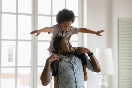 Close up little boy sitting on African American father shoulders, pretending flying with arms outstretched, happy dad and son playing funny active game, family enjoying leisure time together