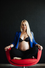 Obraz premium Pregnant woman in a blue jacket and pants sitting in a stylish red chair on a background of black wall. Beautiful pregnant blonde enjoys pregnancy. Nine months.