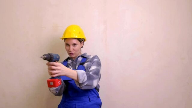 A young, beautiful female construction worker in a yellow hard hat and blue uniform stands, picks up a screwdriver and pretends to shoot it. Girl repairman