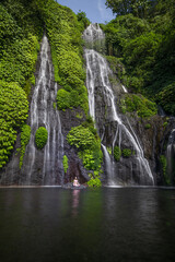 Fototapeta premium Yoga lotus pose. Young Caucasian woman sitting on the stone, meditating, practicing yoga, pranayama at waterfall. Hands in gyan mudra. Yoga retreat. Copy space. Banyumala waterfall, Bali