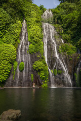 Yoga lotus pose. Young Caucasian woman sitting on the stone, meditating, practicing yoga, pranayama at waterfall. Hands raised up in namaste mudra. Yoga retreat. Banyumala waterfall. Copy space