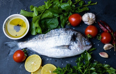 Ready to cook raw sea bream or dorado sea fish. Top view of arugula, lemon, tomato, parsley, garlic, oil and spices on dark background.Mediterranean cuisine, seafood, healthy nutrition.
