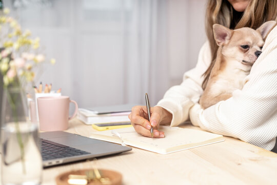 Distance learning online education and work. Business woman with puppy dog writes goals, plans, make to do and wish list in notebook on desk, working from home office. Using laptop.