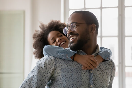 Close up happy African American father wearing glasses piggy backing little daughter, engaged in funny activity, standing at home, cute girl embracing overjoyed dad, family having fun together