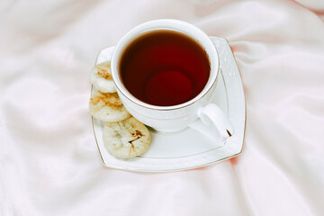 White cup with tea with a saucer, next to dried dates dessert on a silk tablecloth.