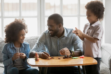 Happy African American father and kids playing with colorful plasticine together, sitting on couch at home, smiling dad wearing glasses with little daughter and son involved in creative activity