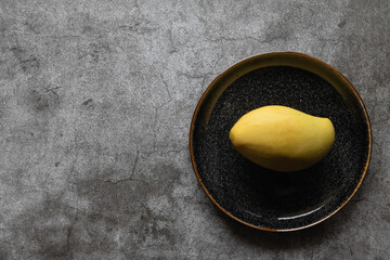 One yellow mango in a dark plate on a concrete background. Copy space, top view. Tropical fruit 