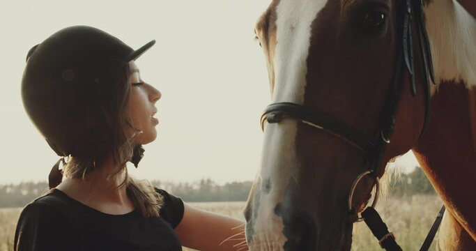 Teen age girl makes preparation for horse riding, putting a bridle on horse. Cute cheerful girl looks at the horse, brown horse with white pigtails, summer field background, non urban scene.
