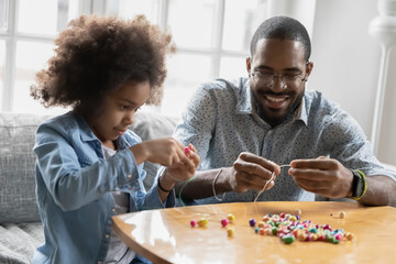Close up smiling African American father and little daughter playing with colorful beads, sitting on couch at home, crafting bracelet, family involved in creative activity, enjoying leisure time
