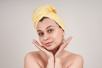 A cute young woman with bare shoulders, a yellow towel on her head, applies a cream to her face. isolate on a gray background.