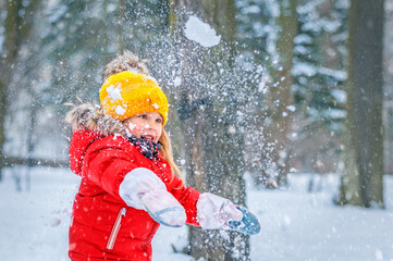 A little girl in a red jacket and a yellow hat laughs in the winter forest and plays with the snow.