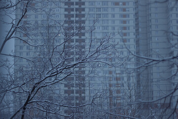 Snow falls. Close-up of silhouettes of trees. In the background is an urban apartment building.