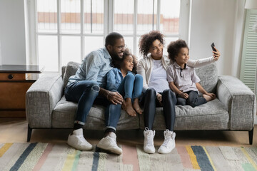 Happy African American family posing for selfie, using phone, little son holding smartphone, photographing, smiling mother and father with adorable son and daughter sitting on couch at home